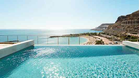 Näkymä infinity-altaasta, jossa veden pinnalla aurinko kimmeltää, ja taustalla hiekkaranta. (Tekoälyn luoma teksti)