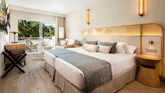 Hotellihuone, jossa on suuri parivuode, istuinryhmä ja parveke näkymällä vihreisiin palmuihin. (Tekoälyn luoma teksti)