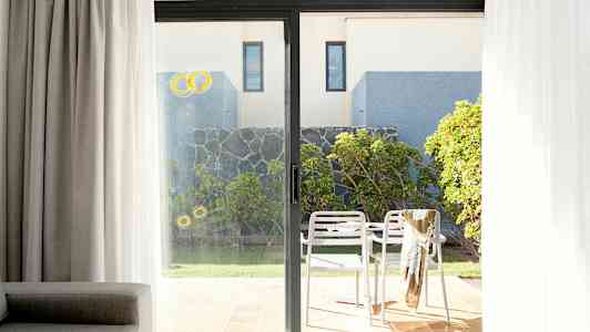 Kuva patioalueesta, jossa kaksi valkoista tuolia ja pöytä, ympärillä kasveja ja kivimuuri, nähtynä lasiseinän läpi. (Tekoälyn luoma teksti)