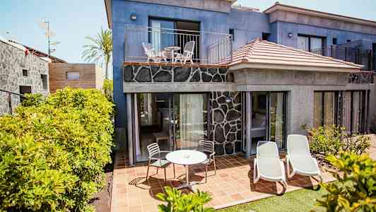 Kuvassa on hotellirakennus, jossa sininen julkisivu ja kivipaneelit. Alimmassa kerroksessa on terassi ulkokalusteilla ja aurinkotuoleilla, jota ympäröivät kasvit. (Tekoälyn luoma teksti)