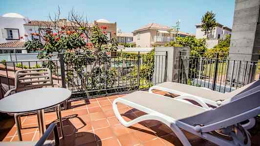 Kuvassa on parveke, jossa on pöytä, kaksi tuolia ja kaksi aurinkotuolia kaiteen edessä, näkymä sisäpihalle ja ympäröiviin rakennuksiin. (Tekoälyn luoma teksti)