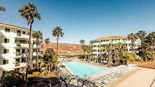 Kuva hotellialueesta, jossa on suuri ulkouima-allas aurinkotuolien ympäröimänä. Kaksi valkoista rakennusta, joissa on vihreät ikkunat, ja etualalla palmuja. (Tekoälyn luoma teksti)