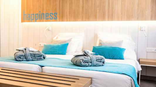 Hotellihuone, jossa on parivuode, sinisiä yksityiskohtia kuten tyynyjä ja päiväpeite sekä sängylle asetetut rullatut pyyhkeet. (Tekoälyn luoma teksti)