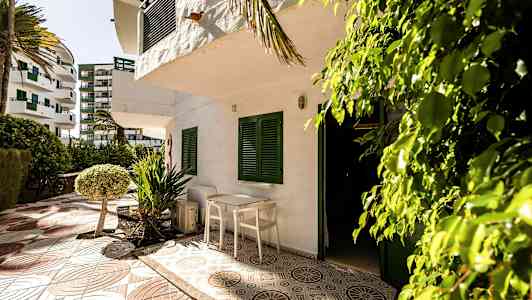 Terassi, jossa on pöytä ja tuolit valkoisen rakennuksen edessä, jossa on vihreät ikkunaluukut. Ympärillä on puita ja pensaita. (Tekoälyn luoma teksti)