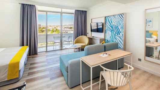 Hotellihuone, jossa on sänky, kirjoituspöytä, sohva, televisio ja lasiovet, jotka johtavat parvekkeelle. (Tekoälyn luoma teksti)