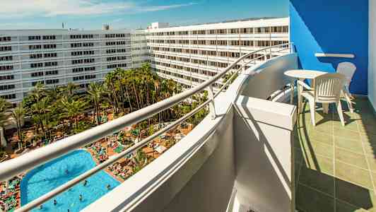 Näköala parvekkeelta uima-allasalueelle, jossa on paljon aurinkotuoleja ja vihreää ympäristöä, valkoisten hotellirakennusten ympäröimänä. (Tekoälyn luoma teksti)