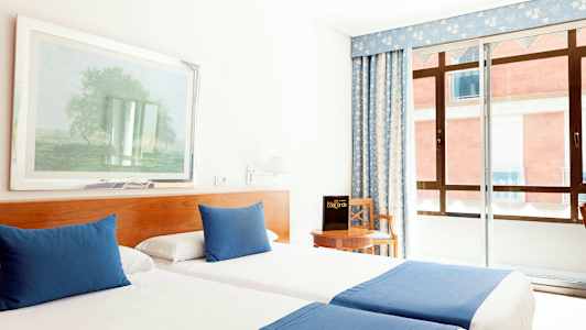 Hotellihuone, jossa on kaksi sinisillä peitoilla ja tyynyillä varustettua yhden hengen sänkyä, tuoli pöydän ääressä ja ikkuna sinisellä verholla. (Tekoälyn luoma teksti)