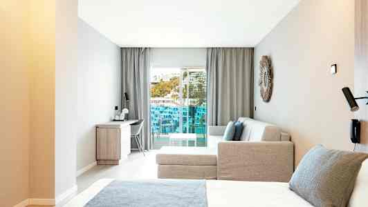 Moderni hotellihuone, jossa on sänky, sohva ja työpöytä sekä parveke, jolta on näköala. (Tekoälyn luoma teksti)