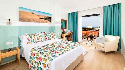 Hotellihuone, jossa on koristeellinen päiväpeite, nojatuoli ja kirjoituspöytä sekä näkymä parvekkeelta. (Tekoälyn luoma teksti)