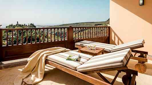 Kaksi raidallisilla tyynyillä varustettua aurinkotuolia seisoo terassilla, josta avautuu näkymä vehreille kukkuloille. (Tekoälyn luoma teksti)