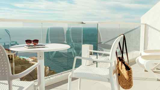 Terassi, jossa on valkoisia huonekaluja, olkilaukku ja näkymä merelle. (Tekoälyn luoma teksti)