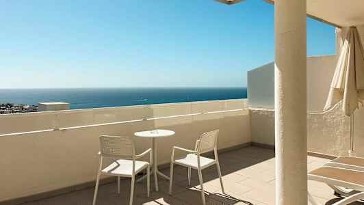 Terassi, jossa kaksi valkoista tuolia ja pöytä, taustalla näkyy meri. (Tekoälyn luoma teksti)