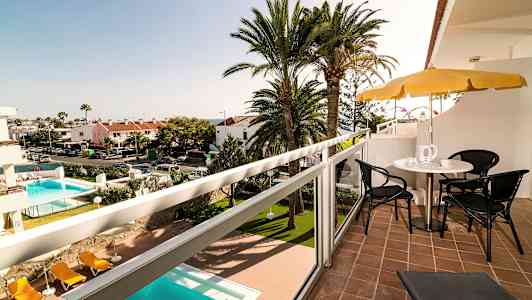 Näkymä parvekkeelta, jossa on istuimet ja aurinkovarjo, taustalla uima-allas ja palmuja. (Tekoälyn luoma teksti)