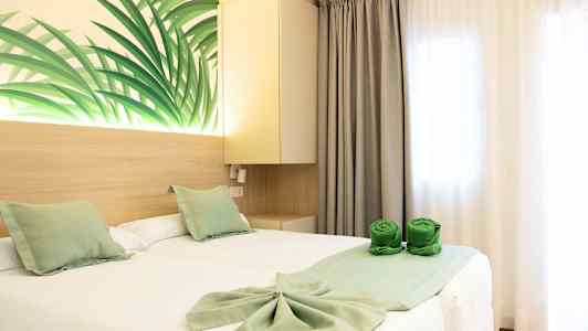 Hotellihuone, jossa on kaksi yhden hengen vuodetta valkoisilla lakanoilla ja vihreillä koristetyynyillä. Seinäkoristeena lehtikuvio sekä vihreät rullatut pyyhkeet sängyllä. (Tekoälyn luoma teksti)