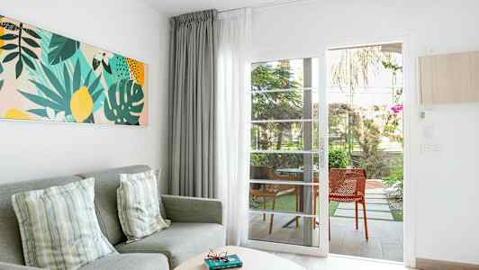 Olohuoneen ympäristö, jossa harmaa sohva, koristetyynyt, värikäs seinätaide ja lasiovi, joka johtaa terassille. (Tekoälyn luoma teksti)