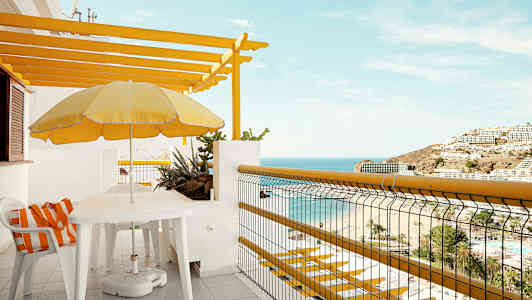 Parveke, josta on näköala merelle ja rannalle, varustettu pöydällä, tuoleilla ja keltaisella aurinkovarjolla. (Tekoälyn luoma teksti)