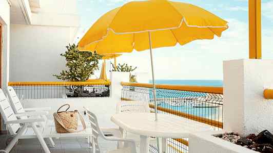Valkoinen terassi, jossa on tuolit, pöytä, keltainen aurinkovarjo ja merinäkymä. (Tekoälyn luoma teksti)