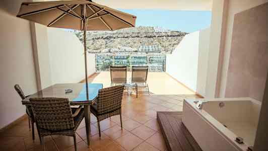 Terassi, jossa ruokapöytä, aurinkovarjo, aurinkotuoleja ja poreallas, näkymä ympäristöön. (Tekoälyn luoma teksti)