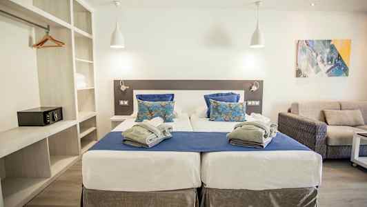 Kahden hengen huone, jossa on kaksi yhden hengen vuodetta, siniset ja valkoiset vuodevaatteet sekä harmaa viltti. Moderni sisustus sohvalla ja hyvin valaistu tunnelma. (Tekoälyn luoma teksti)