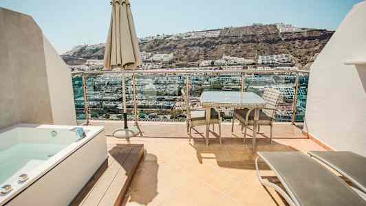 Terassi, jossa on ulkokalusteita, aurinkovarjo ja poreallas, sekä näkymä rinteeseen, jossa rakennuksia. (Tekoälyn luoma teksti)