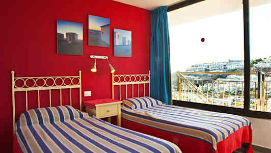 Kaksi yhden hengen sänkyä raidallisilla vuodevaatteilla ja punainen seinä, jossa on kuvia, sekä suuri ikkuna näkymällä rakennuksiin. (Tekoälyn luoma teksti)