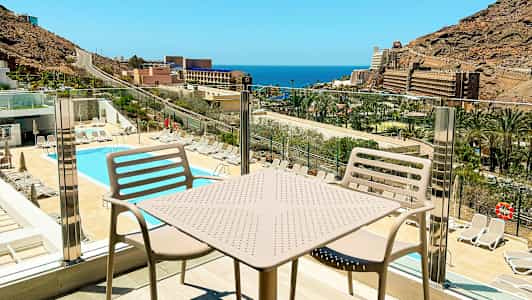 Näkymä uima-altaalle ja aurinkotuoleihin, taustalla meri ja rakennuksia, nähtynä parvekkeelta, jossa on pöytä ja tuolit. (Tekoälyn luoma teksti)