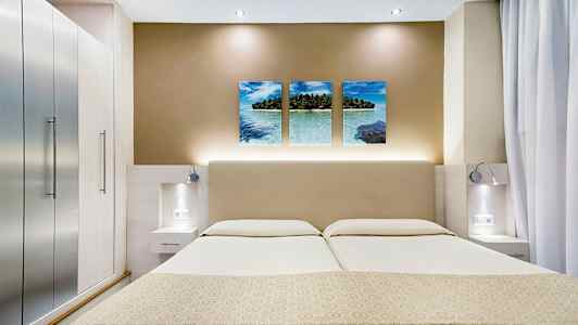 Kuvassa on moderni hotellihuone, jossa on parivuode, tyylikkäät yöpöydät ja seinäkoriste, johon kuuluu kolme trooppista rannalla tehtyä kuvaa. (Tekoälyn luoma teksti)