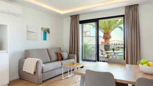 Olohuone, jossa on harmaa sohva, sohvapöytä ja ruokapöytä. Ulkona näkyy terassi ulkokalusteineen ja palmupuu. (Tekoälyn luoma teksti)