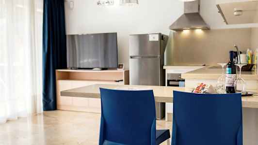 Keittiö, jossa tiskipöytä, kaksi sinistä tuolia, jääkaappi ja televisio puukaapin luona. (Tekoälyn luoma teksti)