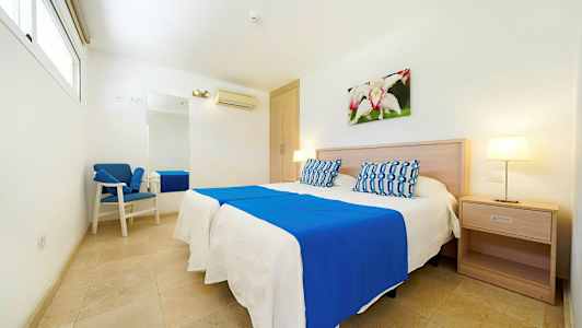 Valoisa hotellihuone, jossa on parivuode, siniset ja valkoiset vuodevaatteet, yöpöytä, seinäkoriste ja nojatuoli. (Tekoälyn luoma teksti)