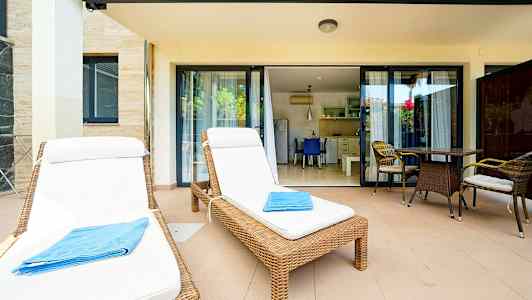 Kaksi aurinkotuolia valkoisilla tyynyillä ja sininen pyyhe terassilla, jossa on pöytä ja tuolit lasiovien edessä keittiöön. (Tekoälyn luoma teksti)