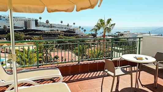 Terassi, jossa on pöytä, tuolit ja aurinkotuolit. Taustalla näkyy palmuja, rakennuksia ja meri. (Tekoälyn luoma teksti)