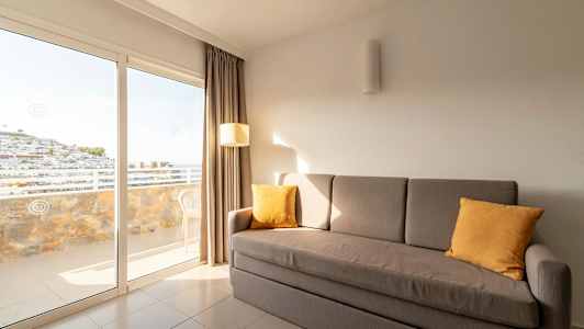 Harmaa sohva, jossa on keltaisia ​​tyynyjä, seisoo huoneessa, jossa suuret lasiovet avautuvat parvekkeelle. (Tekoälyn luoma teksti)