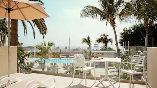 Parveke, jossa aurinkotuoleja ja pöytä tuoleineen, näkymä uima-altaalle ja palmuihin. (Tekoälyn luoma teksti)