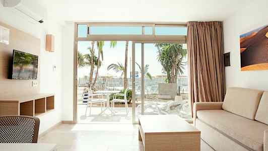 Valoisa huone, jossa on sohva, pöytä ja televisio sekä näkymä terassille, jolla on pöytä ja tuolit. Ulkona näkyy palmupuita. (Tekoälyn luoma teksti)