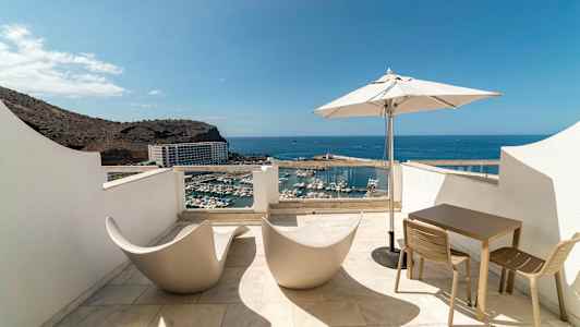 Terassi, jolta on näkymä satamaan ja merelle, varustettuna moderneilla tuoleilla, pöydällä ja aurinkovarjolla. (Tekoälyn luoma teksti)