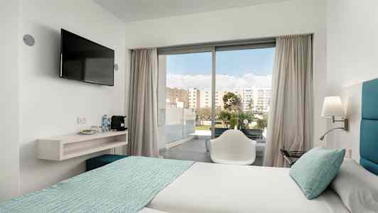 Hotellihuone, jossa kaksi vuodetta vaaleilla lakanoilla ja turkooseilla päiväpeitteillä. Tuoli on lasioven edessä, joka johtaa parvekkeelle. Seinään kiinnitetty televisio on hyllyn vieressä, jossa on vesipulloja ja kahvinkeitin. (Tekoälyn luoma teksti)