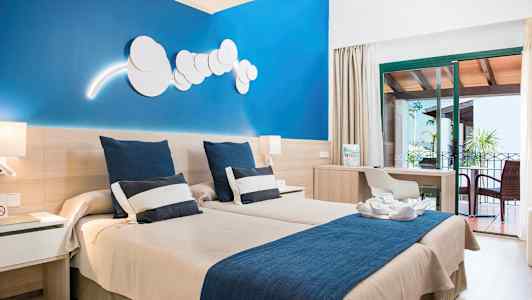 Hotellihuone, jossa kaksi yhden hengen vuodetta, sininen seinä, kirjoituspöytä ja kalustettu parveke. (Tekoälyn luoma teksti)