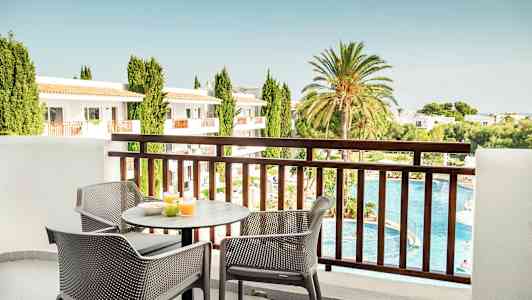 Näkymä parvekkeelta, jossa on pöytä ja tuoleja, joille on laitettu aamiaisia. Taustalla on uima-allas, viheralueita ja rakennuksia. (Tekoälyn luoma teksti)