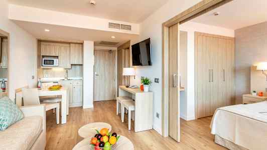 Moderni hotellihuone, jossa on keittiöalue, pöytä, sohva ja makuuhuone. Sohvapöydällä on kulho tuoreita hedelmiä. (Tekoälyn luoma teksti)