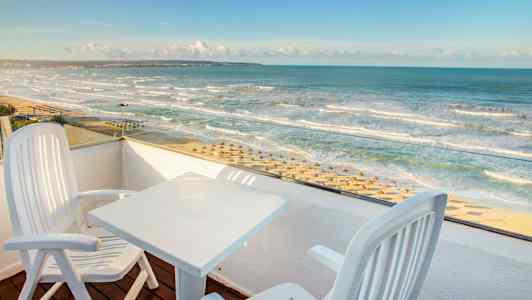 Näkymä parvekkeelta, jossa kaksi tuolia ja pöytä, kohti rantaa, jolla on aurinkovarjoja ja aaltoileva meri. (Tekoälyn luoma teksti)