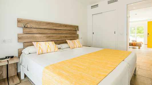 Valoisa makuuhuone, jossa suuri sänky, koristeltu keltaisella päiväpeitteellä ja tyynyillä, sekä puupöytä ja ovi, joka johtaa toiseen huoneeseen. (Tekoälyn luoma teksti)