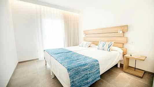 Kaksi yhden hengen sänkyä, joissa valkoiset lakanat ja siniset päiväpeitot, sijoitettu valoisaan ja moderniin huoneeseen, jossa on vieressä puinen pöytä. (Tekoälyn luoma teksti)