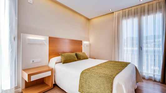 Hotellihuone, jossa on kaksi vihreää tyynyä ja peitto valkoisella sängyllä. Yöpöytä sijaitsee sängyn vieressä. (Tekoälyn luoma teksti)