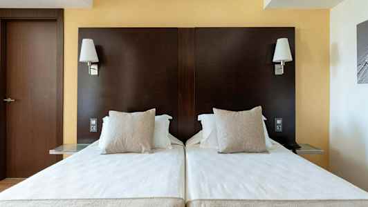 Kaksi yhden hengen sänkyä, joissa valkoiset lakanat ja beiget tyynyt tummanruskeaa sängynpäätyä vasten, seinävalaisimet yläpuolella. (Tekoälyn luoma teksti)