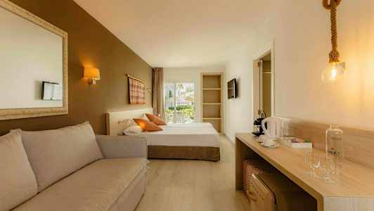 Hotellihuone, jossa sohva, sänky, työpöytä kahvinkeittimen ja laseilla sekä ovi parvekkeelle. (Tekoälyn luoma teksti)