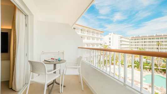 Kalustettu parveke, jossa kaksi tuolia ja pöytä, näkymä hotellialueelle ja uima-altaalle. (Tekoälyn luoma teksti)