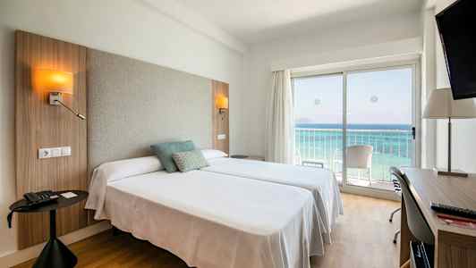 Hotellihuone, jossa on kaksi sänkyä, merinäköala lasioven läpi ja kirjoituspöytä. (Tekoälyn luoma teksti)