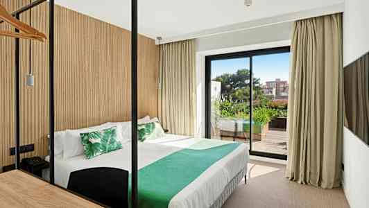 Hotellihuone, jossa vuode on pedattu valkoisilla lakanoilla ja vihreillä tyynyillä, suuri lasiovi johtaa parvekkeelle, jossa on kasvillisuutta. (Tekoälyn luoma teksti)