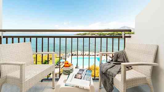 Kaksi valkoista tuolia ja pöytä parvekkeella, josta on näkymä merelle, uima-altaalle ja maisemaan. (Tekoälyn luoma teksti)